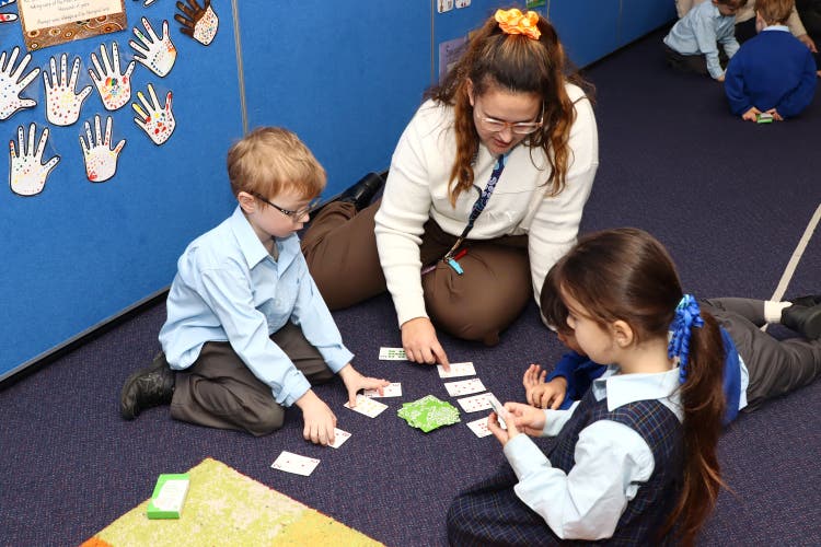 A teacher sitting on the floor playing a card game with two Kindergarten students.